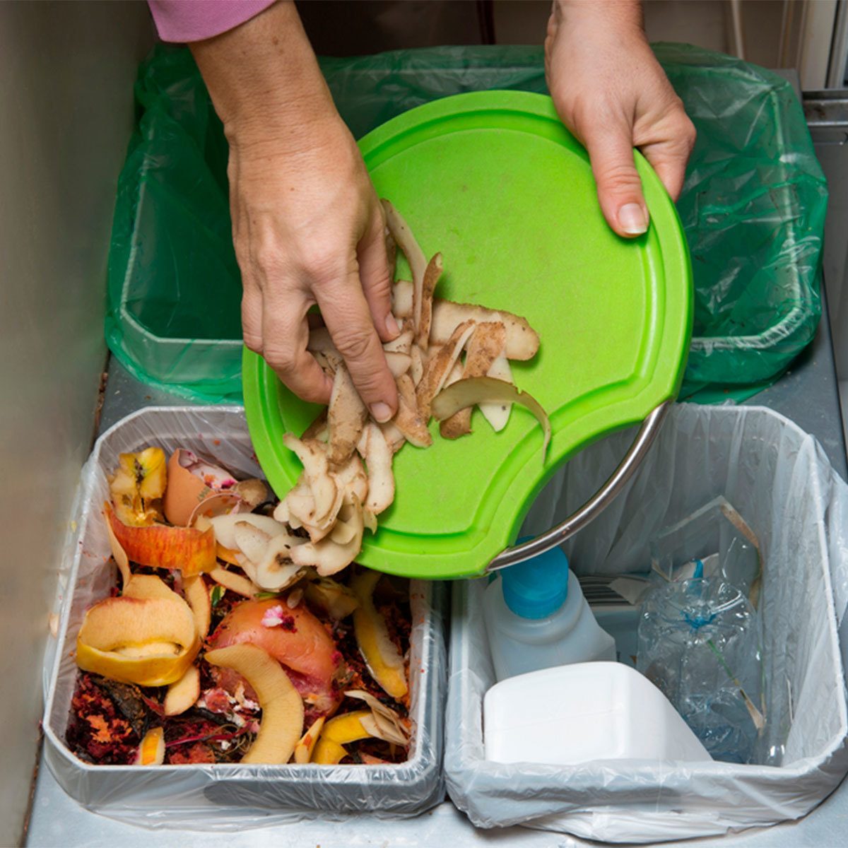 A hand tilts a green cutting board, dumping vegetable peels into a bin filled with organic waste, next to another bin containing plastic and liquid containers.