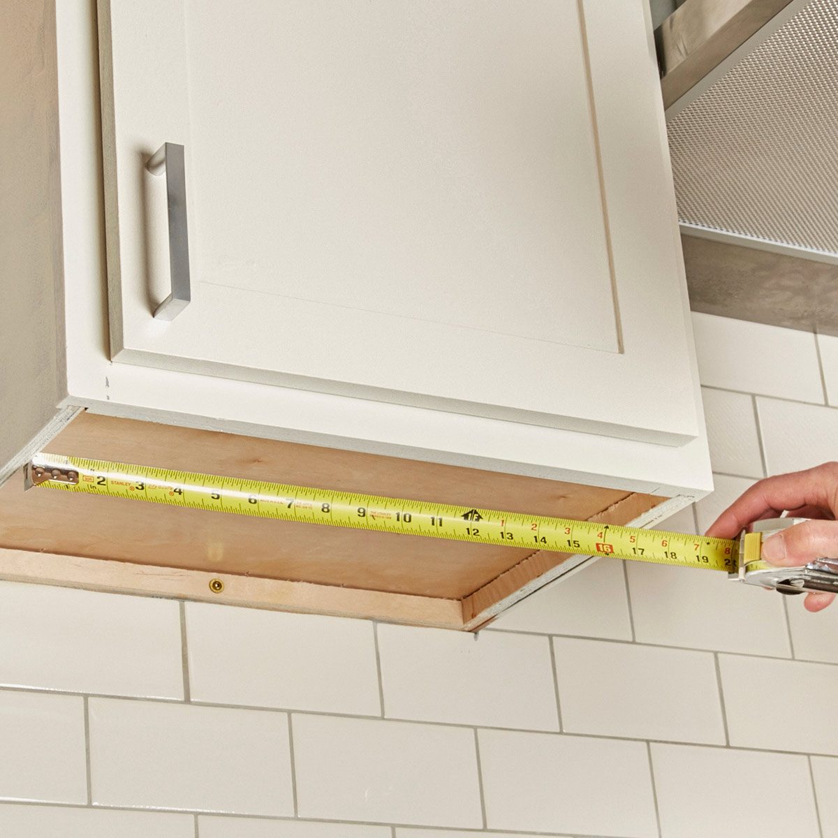 A person's hand measures the interior of a white cabinet with a tape measure, set against a background of white subway tiles.