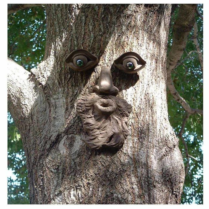 A wooden tree trunk displays a sculpted face, featuring wide eyes, a prominent nose, and a beard, blending artfully with the surrounding natural foliage.