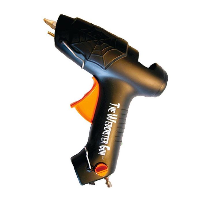 A black and orange tool resembling a gun dispenses adhesive in a workshop or crafting setting, designed for applying glue precisely to various surfaces.