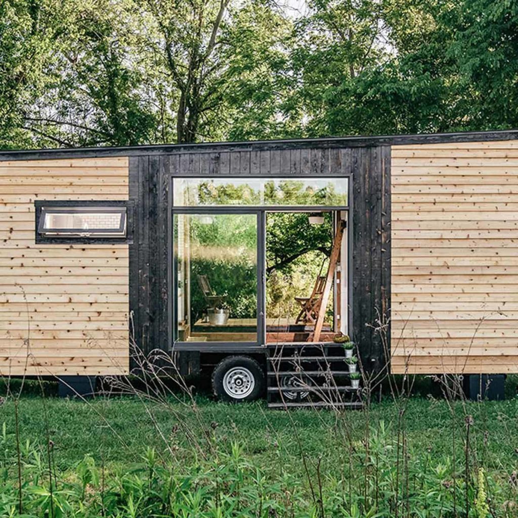 A small, modern wooden cabin features large glass doors, revealing a cozy interior. It sits on a grassy area bordered by lush trees.