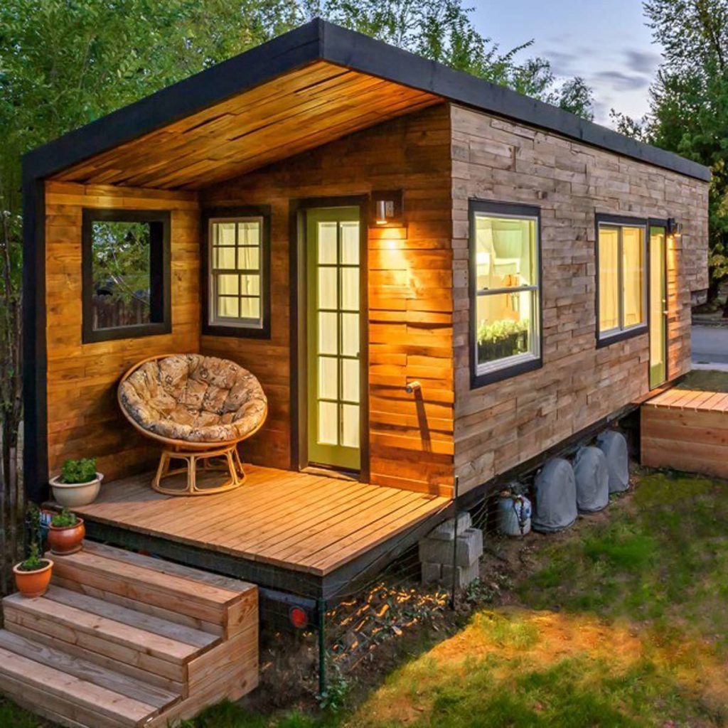 A small wooden house features a welcoming porch with a round chair, illuminated by a nearby light, surrounded by grass and trees.