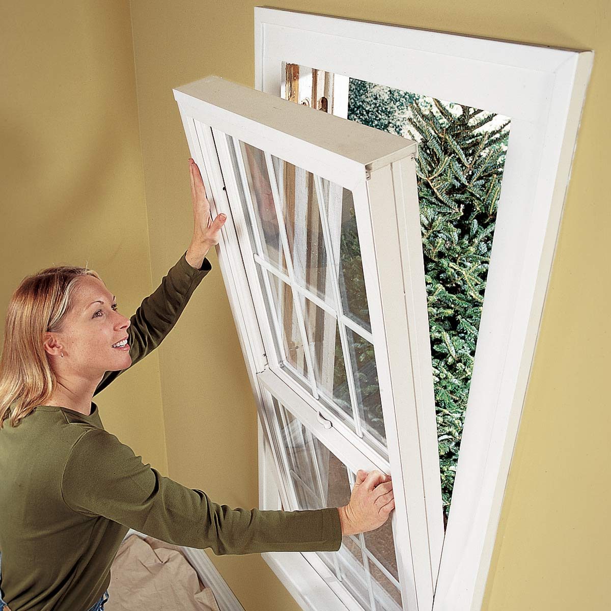 A woman is lifting a partially opened window, revealing a green tree outside. The room has yellow walls and a covered floor.