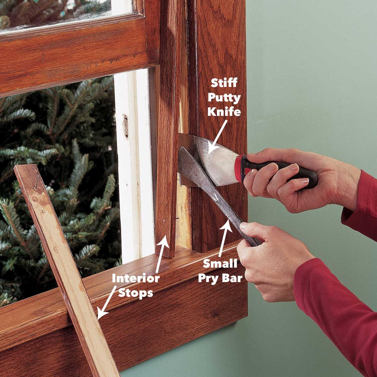 A person uses a stiff putty knife and a small pry bar to remove interior stops from a wooden window frame in a room with green walls.