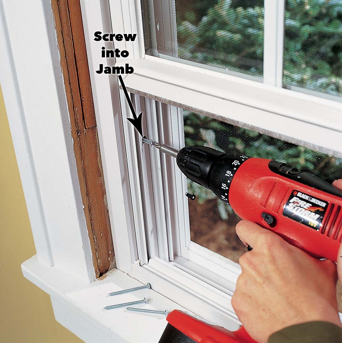 A person uses a cordless drill to screw a fastener into a window jamb, with screws placed nearby on a windowsill.