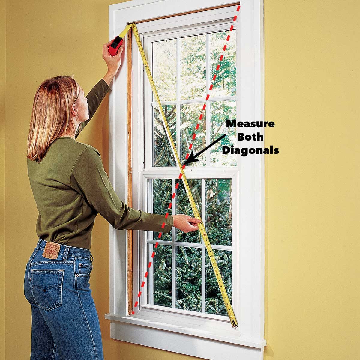 A woman measures a window's diagonals using a tape measure, standing in a room with yellow walls and a view of greenery outside.