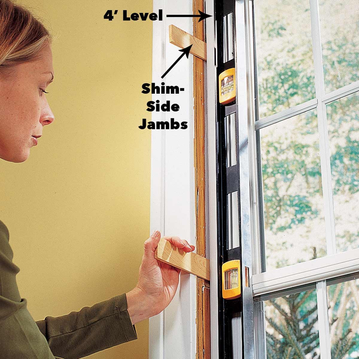 A woman adjusts a shim against a window frame while using a level to ensure it's straight, with a bright yellow wall visible in the background.