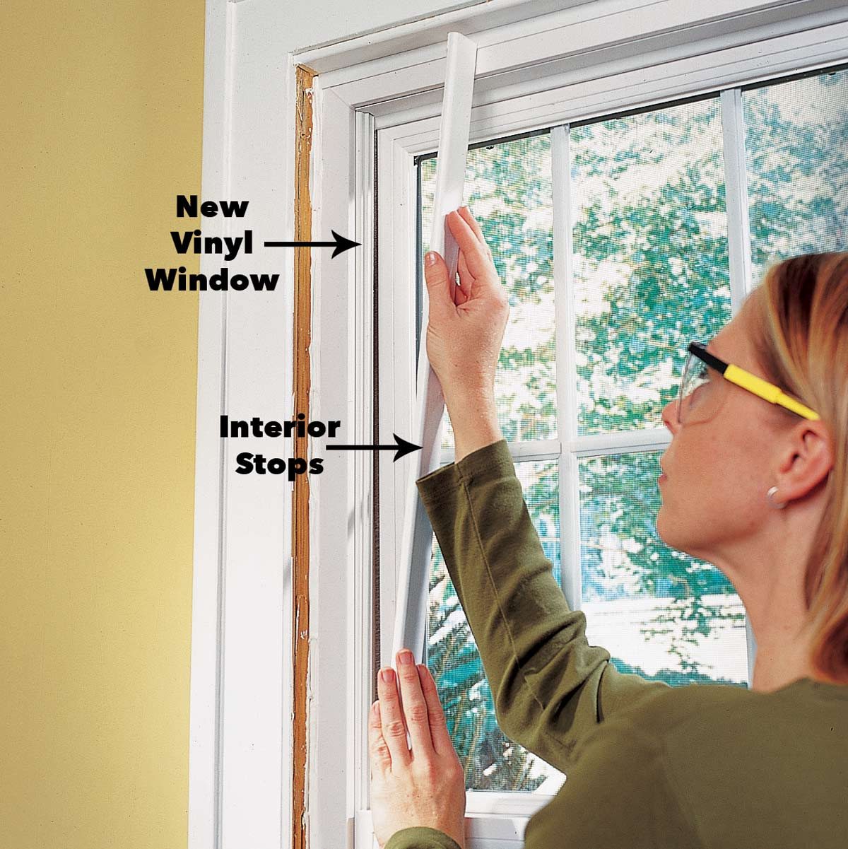 A person installs a new vinyl window, adjusting interior stops, in a room with a large window and yellow walls, revealing greenery outside.