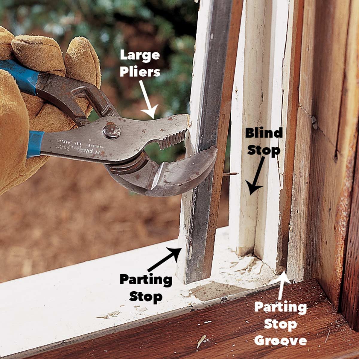 Large pliers grip a parting stop within a window frame, demonstrating the removal process amidst a backdrop of wooden materials and greenery.