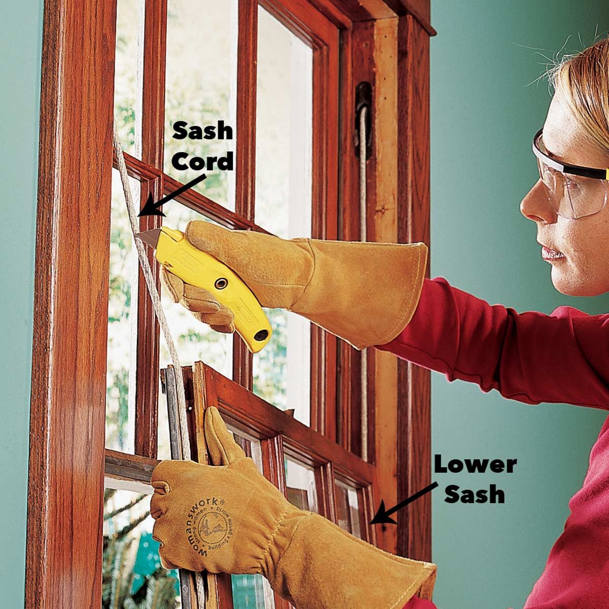 A person in protective gear uses a utility knife to cut a sash cord while working on a wooden window inside a room with blue walls.