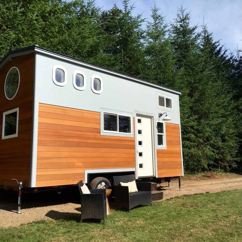 A small, modern, wooden tiny house sits on wheels, surrounded by tall green trees, with two outdoor chairs placed nearby on a gravel patch.
