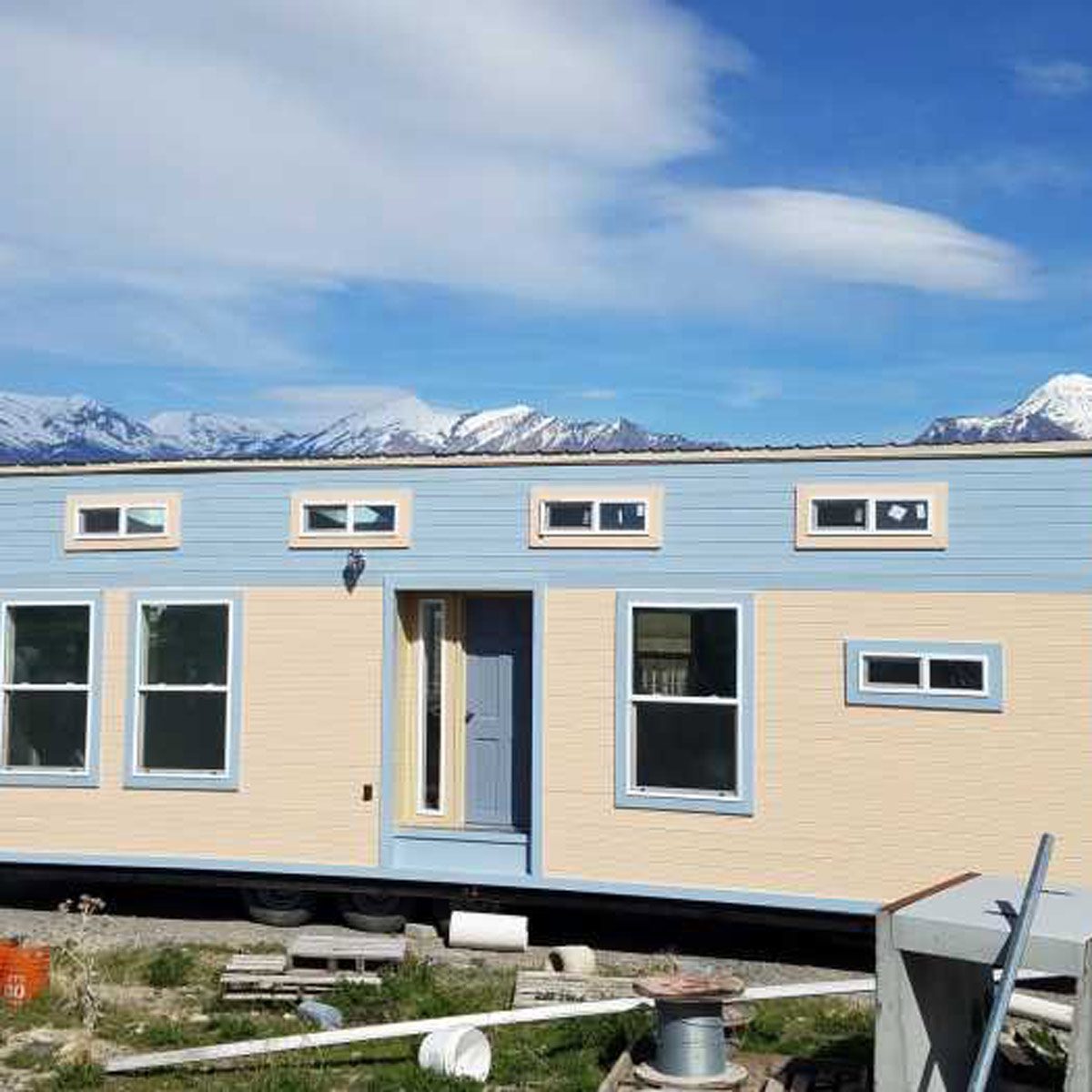 A colorful tiny house on wheels stands outdoors. It features several windows, a front door, and is surrounded by a mountainous landscape under a blue sky.