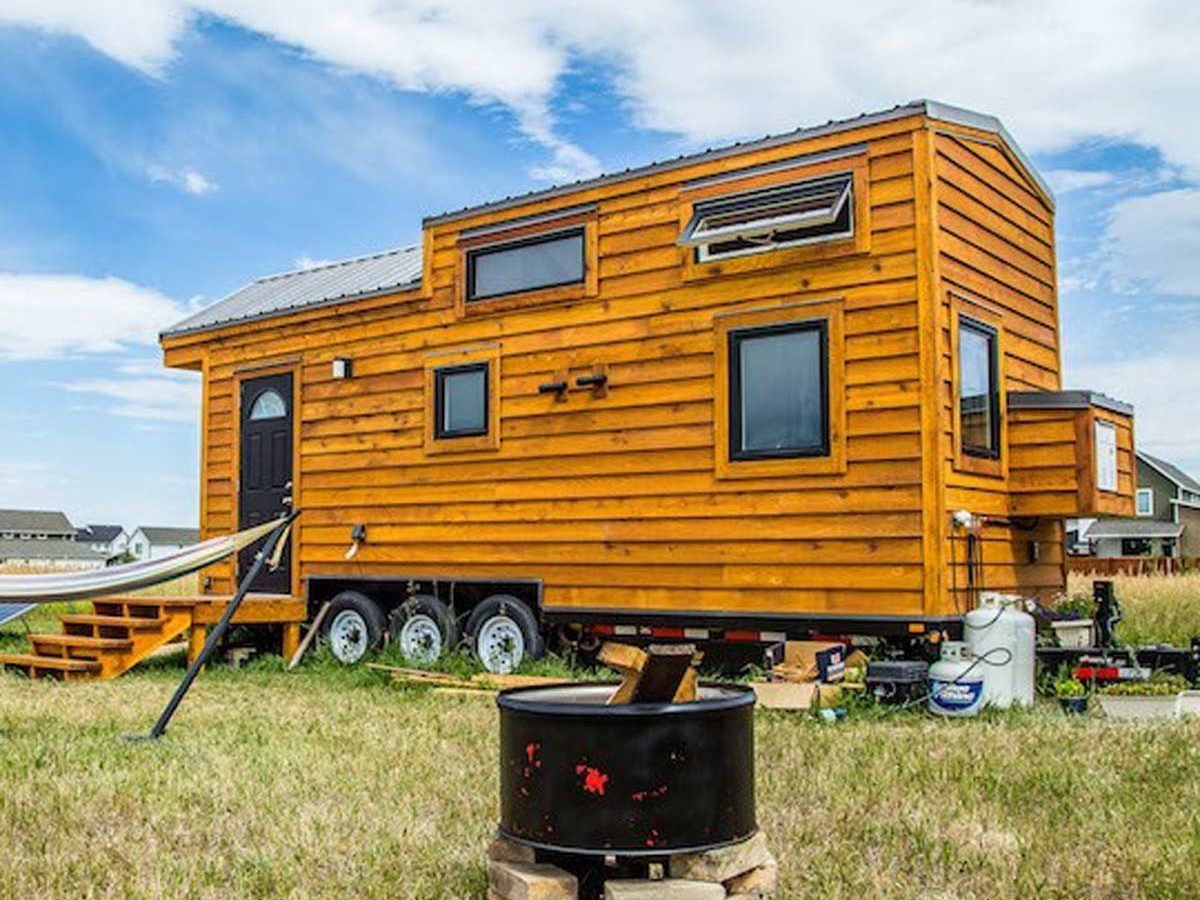A wooden tiny house on wheels is parked on grassy land, with stairs leading to the door, beside a fire pit made of stones.