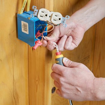 A person connects electrical wires to a blue outlet box mounted on a wooden wall, using a tool in one hand while holding the wires with the other.