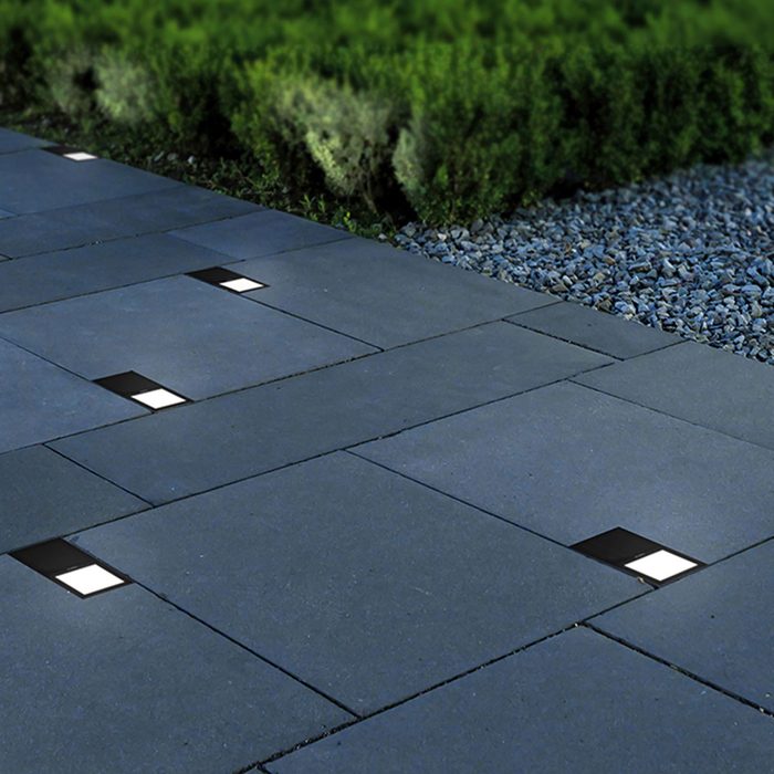 Compact pathway lights embedded in dark stone pavers illuminate the walkway, surrounded by neatly trimmed greenery and a gravel border.