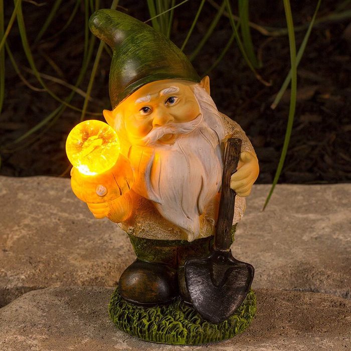 A garden gnome holds a glowing yellow orb in one hand and a small shovel in the other, standing on a stone surface surrounded by grass.