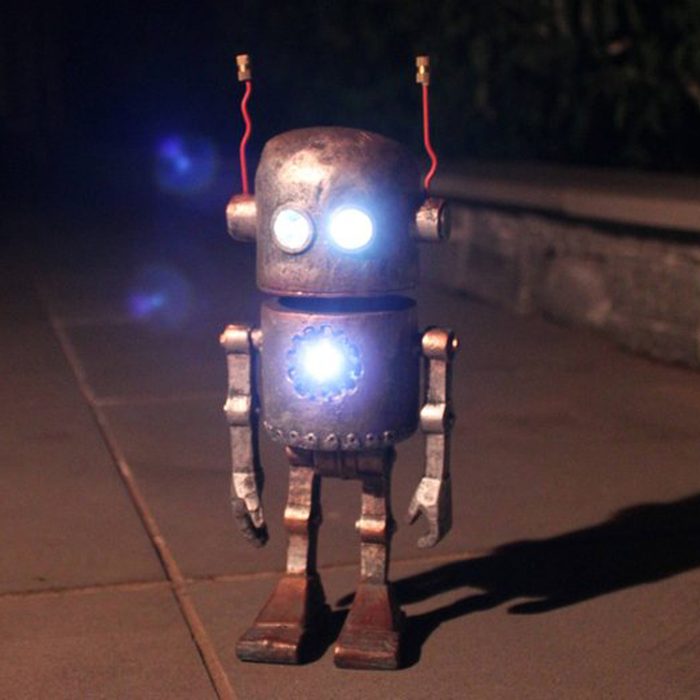 A metallic robot with glowing eyes stands on a stone pathway, illuminated by its own light in a dimly lit outdoor setting.