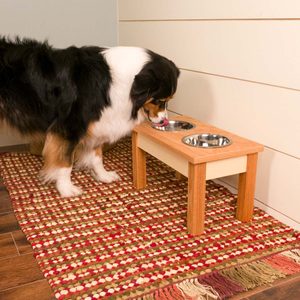 A dog with a black and brown coat is eating from a raised feeding station with two metal bowls, situated on a colorful woven rug.
