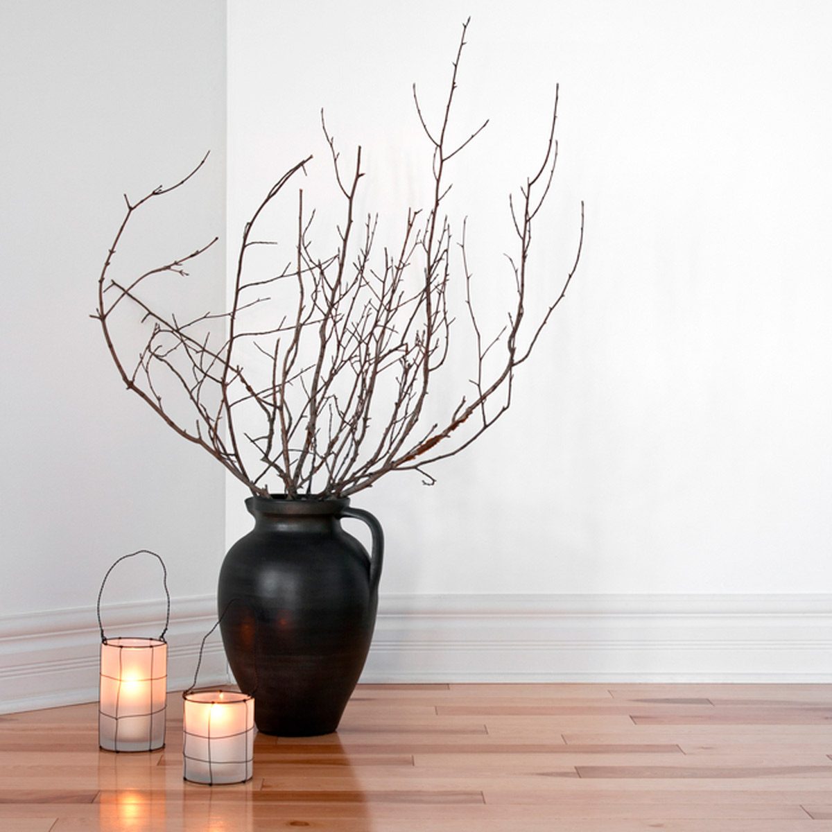 A black vase holds bare branches, standing on polished wooden flooring near two lit candles in wire frames against a bright, minimalistic wall.