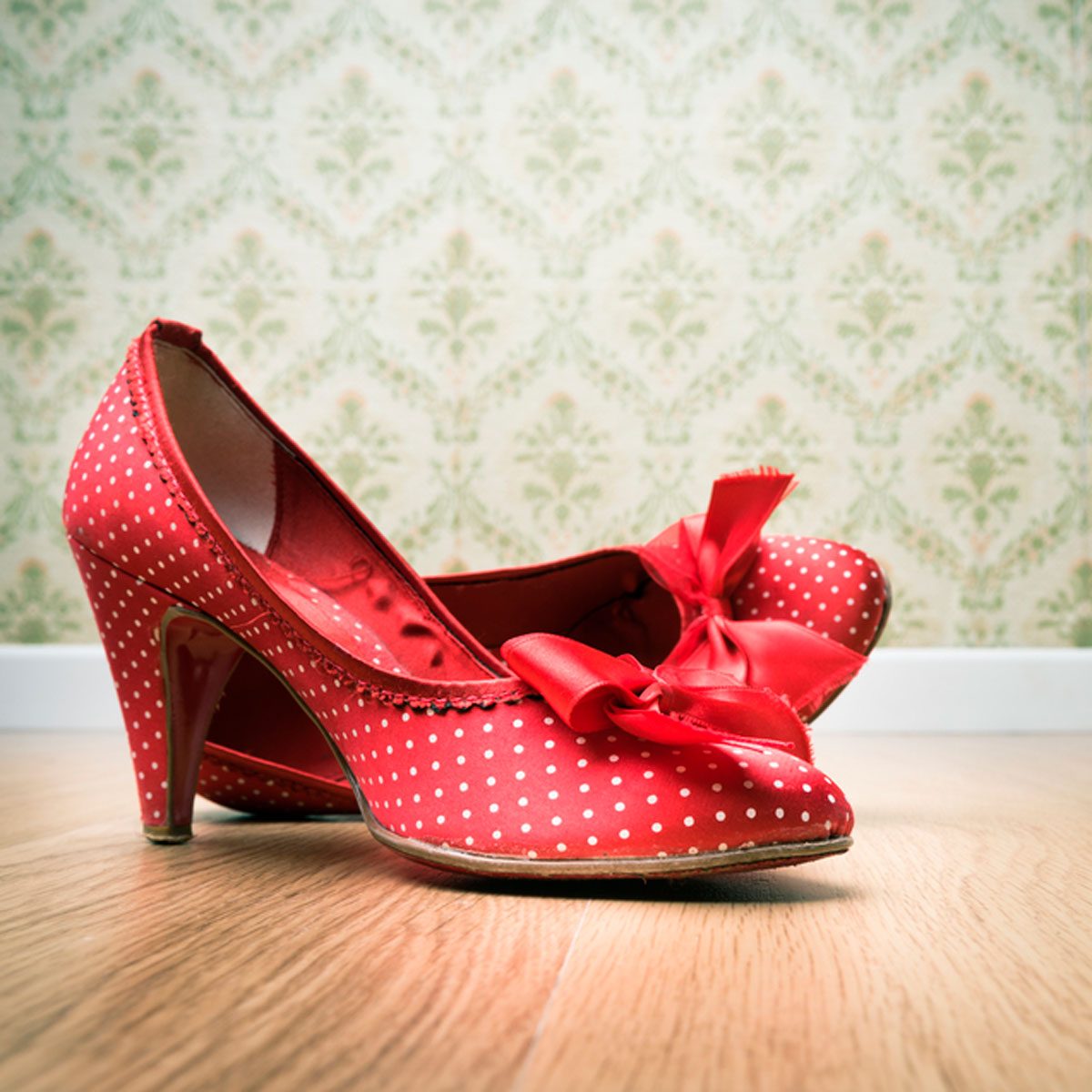 Red polka-dot high heels rest on a wooden floor, with a patterned wallpaper background enhancing their playful and vibrant style.