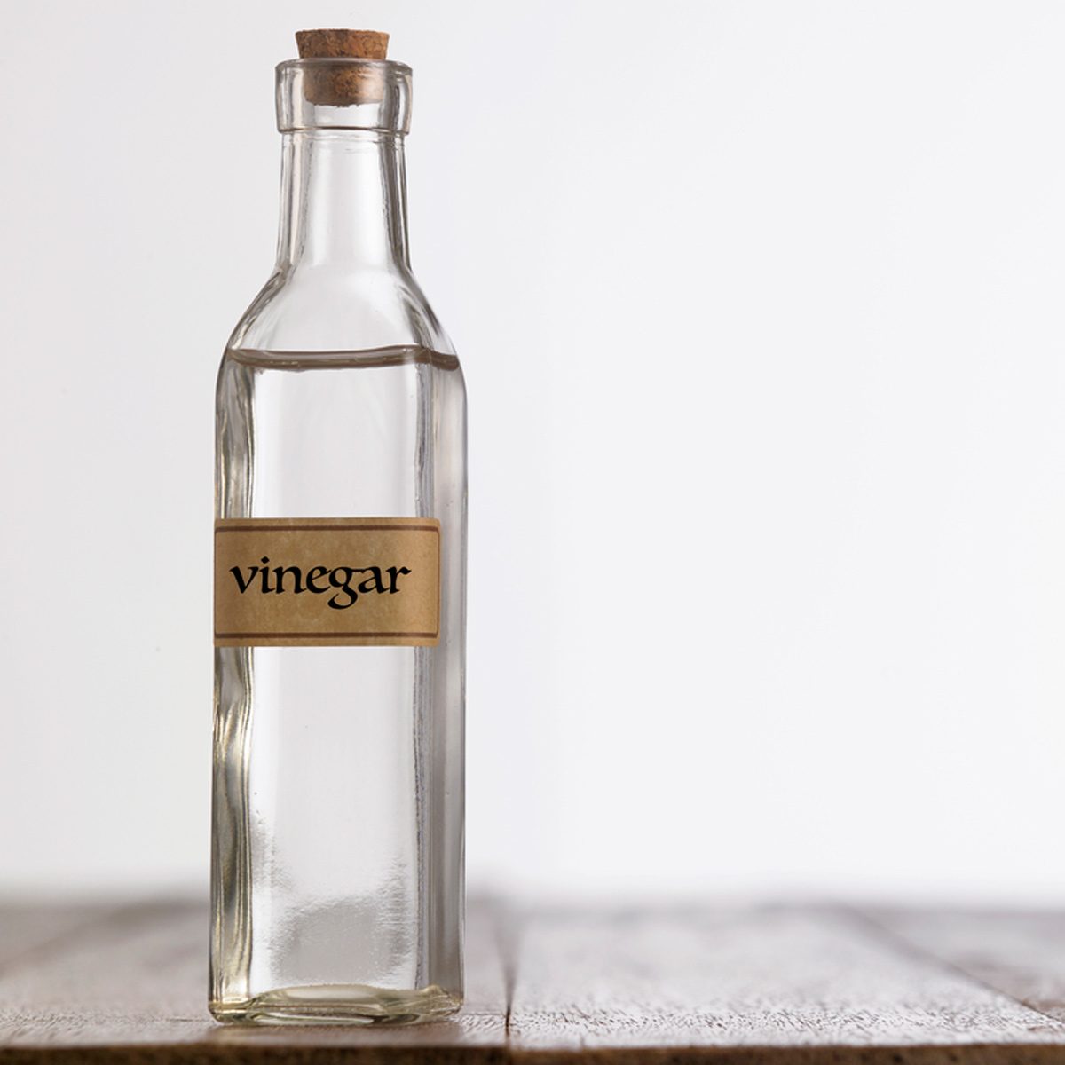 A glass bottle labeled "vinegar" stands upright with a cork stopper on a wooden surface, against a plain, light background.
