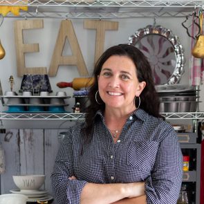 A woman stands smiling with crossed arms in front of colorful kitchen shelves displaying various utensils and decorative items, including the word "EAT."
