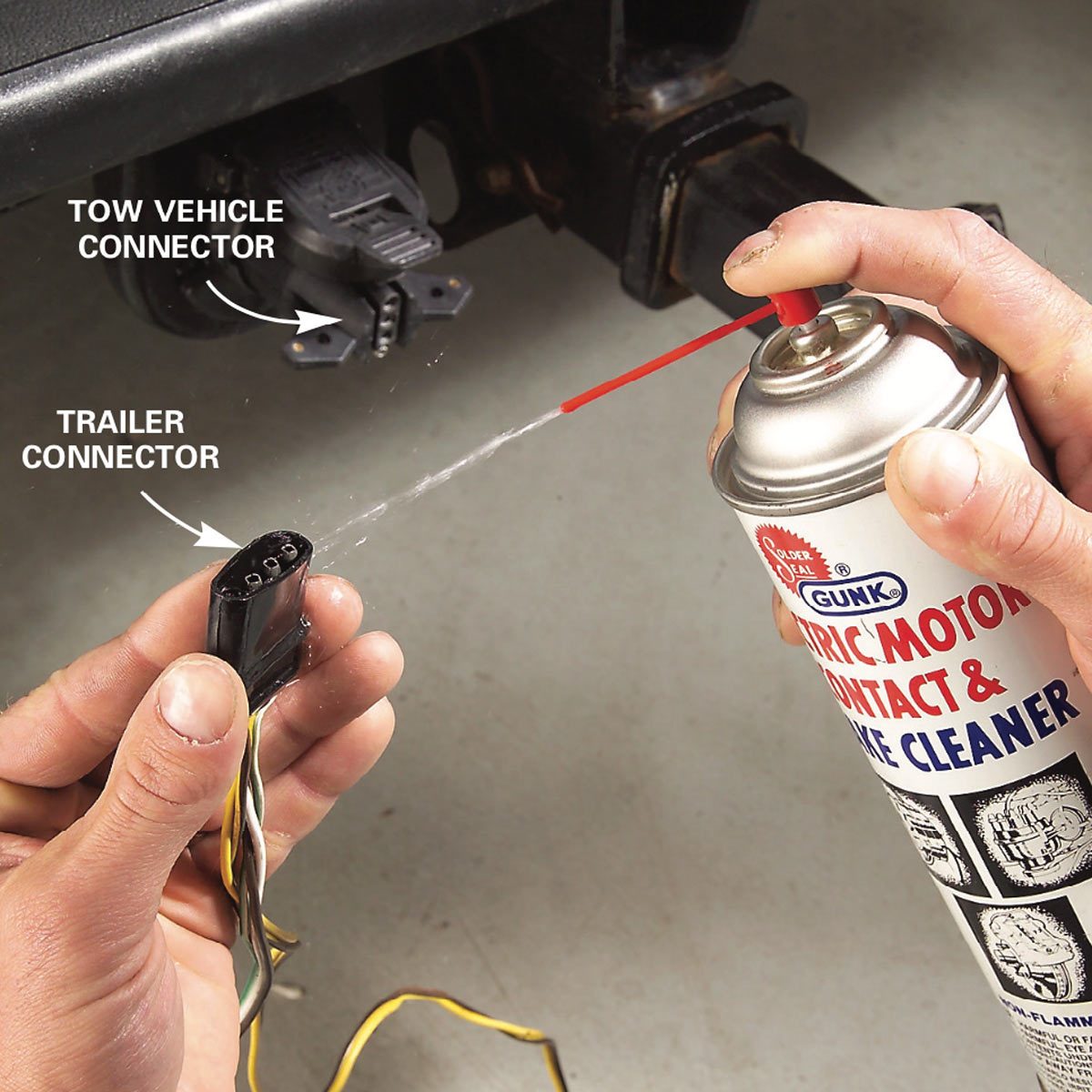 A person sprays cleaner from a can onto a trailer connector while holding it, adjacent to a tow vehicle connector, in a garage setting.