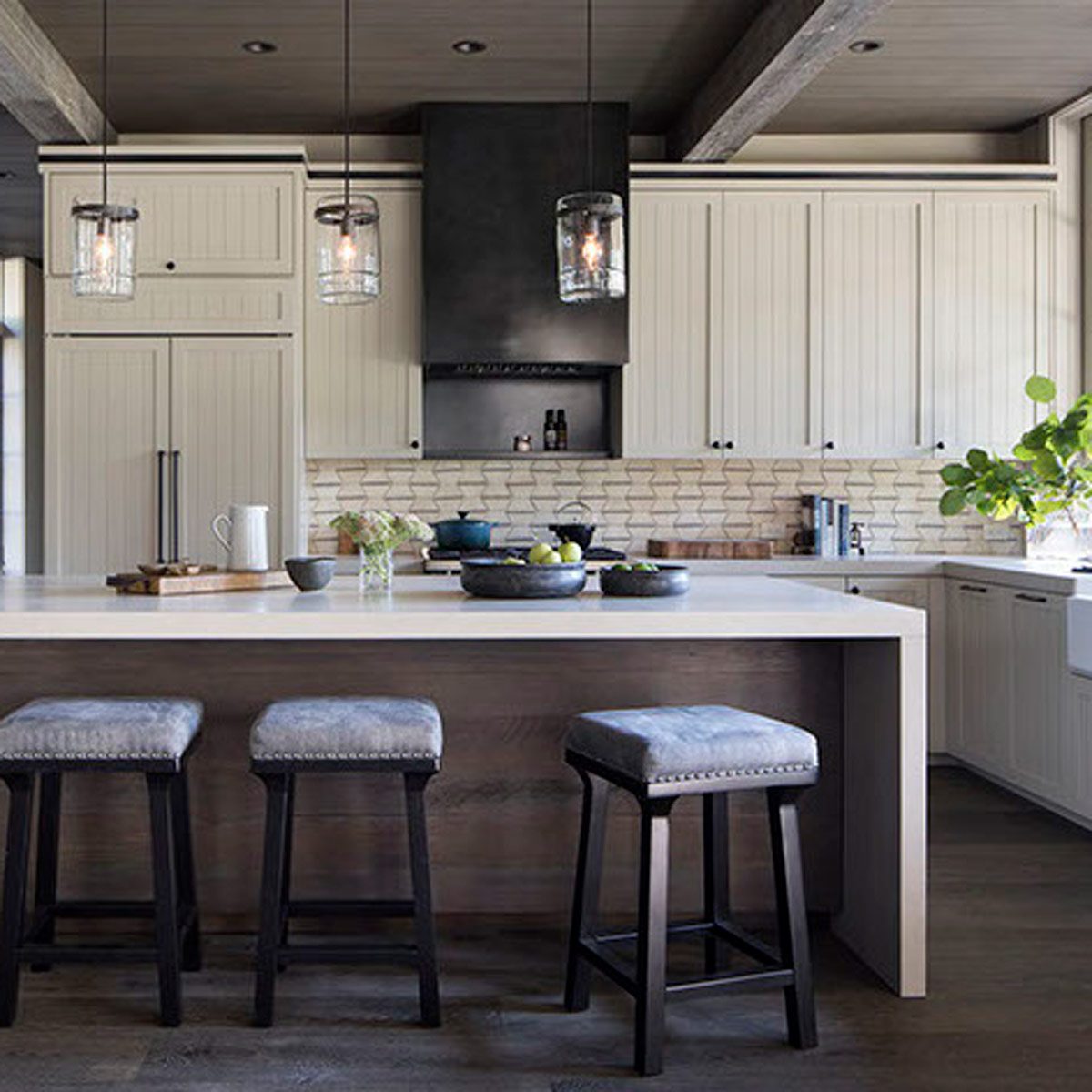 A kitchen island with gray upholstered stools is displayed, featuring a variety of pots, plants, and modern cabinetry under pendant lights in a bright setting.
