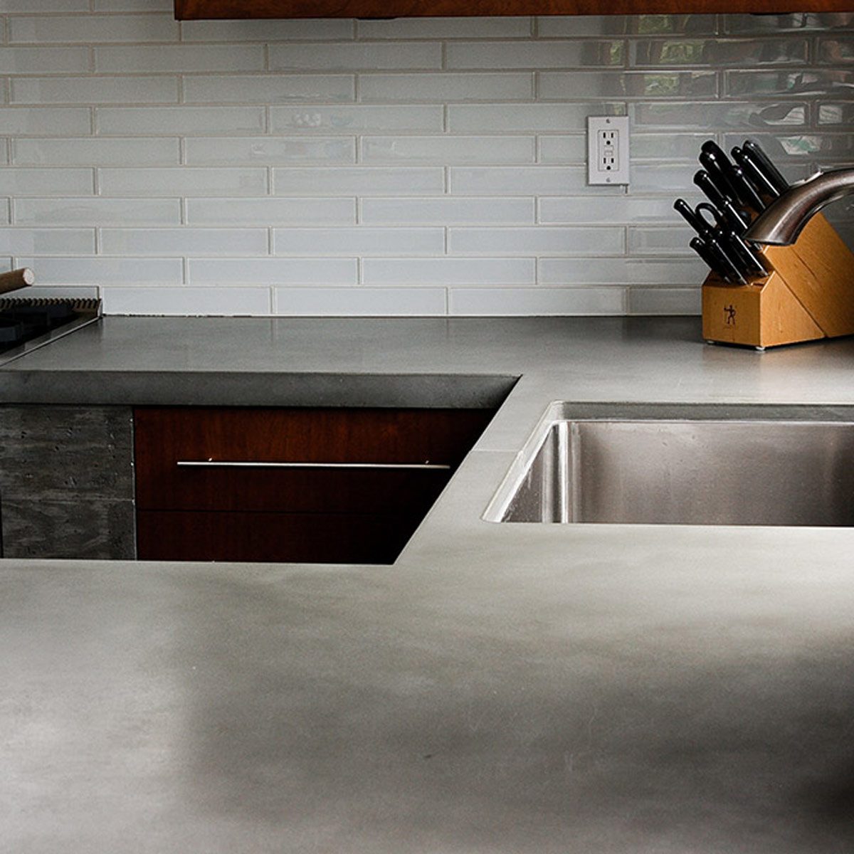 A sleek kitchen countertop features a stainless steel sink, a wooden knife block, and a gas stove, surrounded by light-colored tiled backsplash.