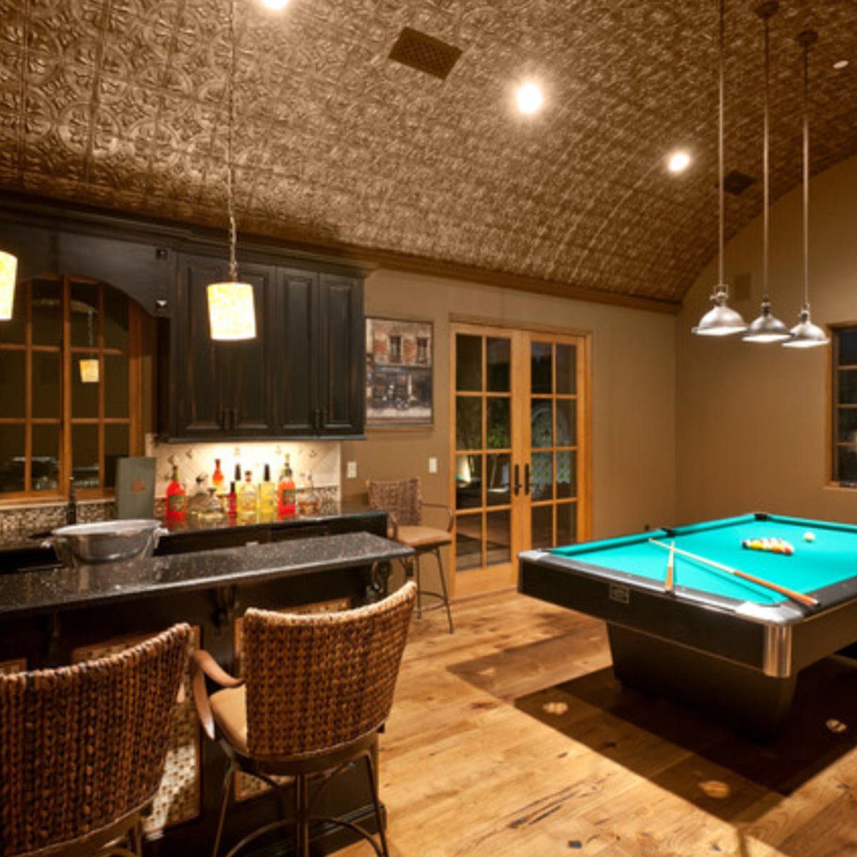 A pool table occupies a spacious room with a cozy bar area, featuring warm lighting and wooden floors, creating an inviting atmosphere for relaxation.