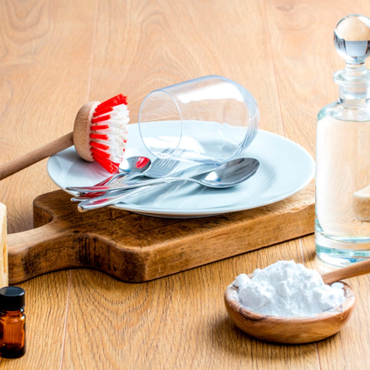 A plate, utensils, and a glass are arranged on a wooden board, alongside a cleaning brush, a glass bottle, and a bowl of powder.