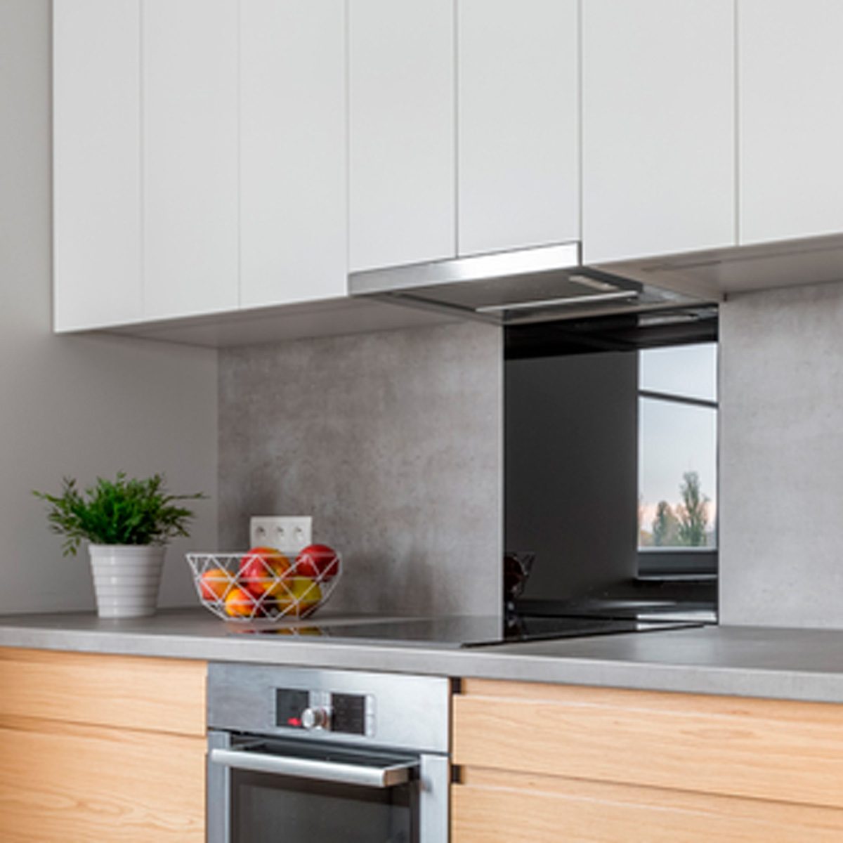 A stainless steel hood hovers above a smooth countertop, where a bowl of colorful fruit rests next to a small potted plant in a modern kitchen setting.