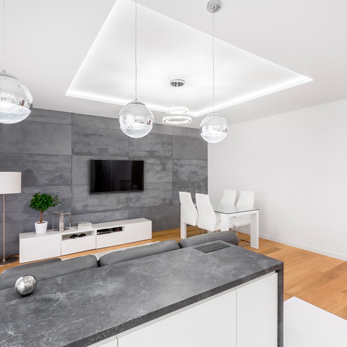 A modern living room features a sectional sofa and sleek coffee table, while pendant lights illuminate the area. A dining table stands nearby against a neutral backdrop.