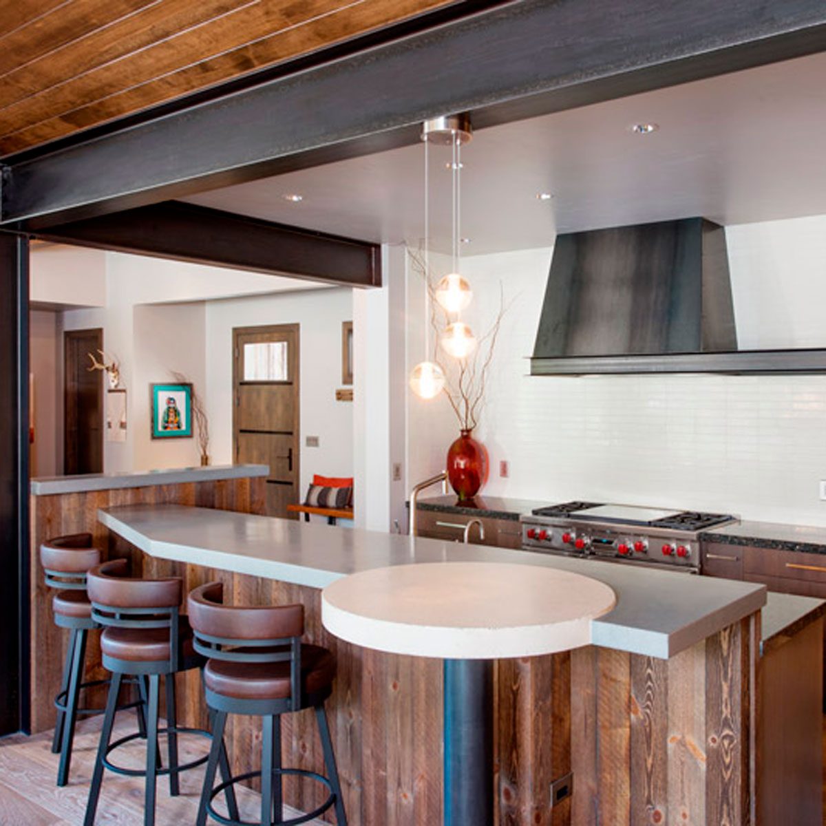 A sleek kitchen features a countertop with four stools, modern lighting, and a stove below a vent. Wood accents enhance the contemporary design.