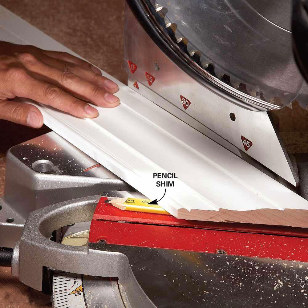 A person's hand holds white trim against a miter saw, with a labeled pencil shim underneath, ready for cutting in a workshop setting.