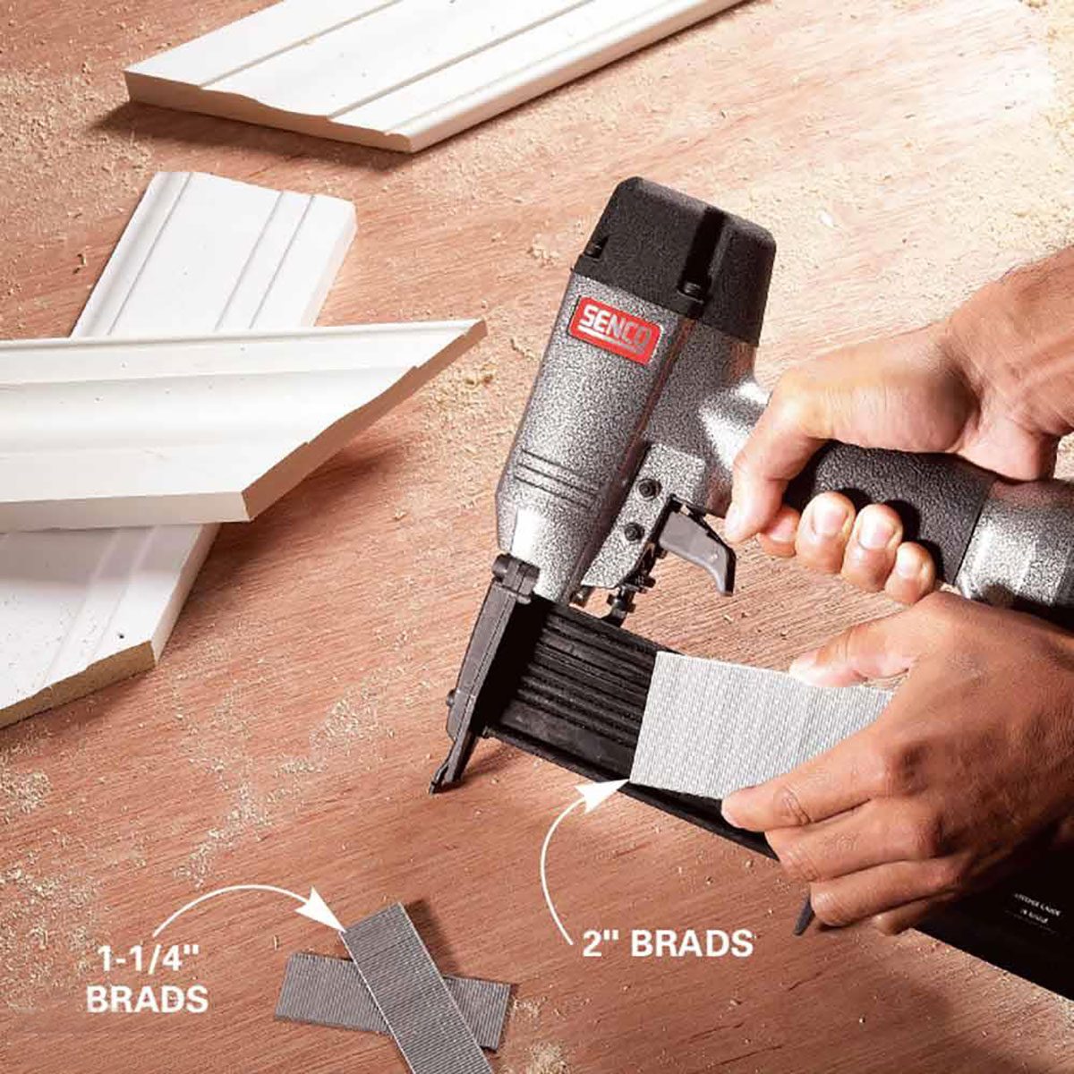 A hand holds a nail gun, loading brads onto a work surface with wooden trim pieces nearby, surrounded by wood shavings.
