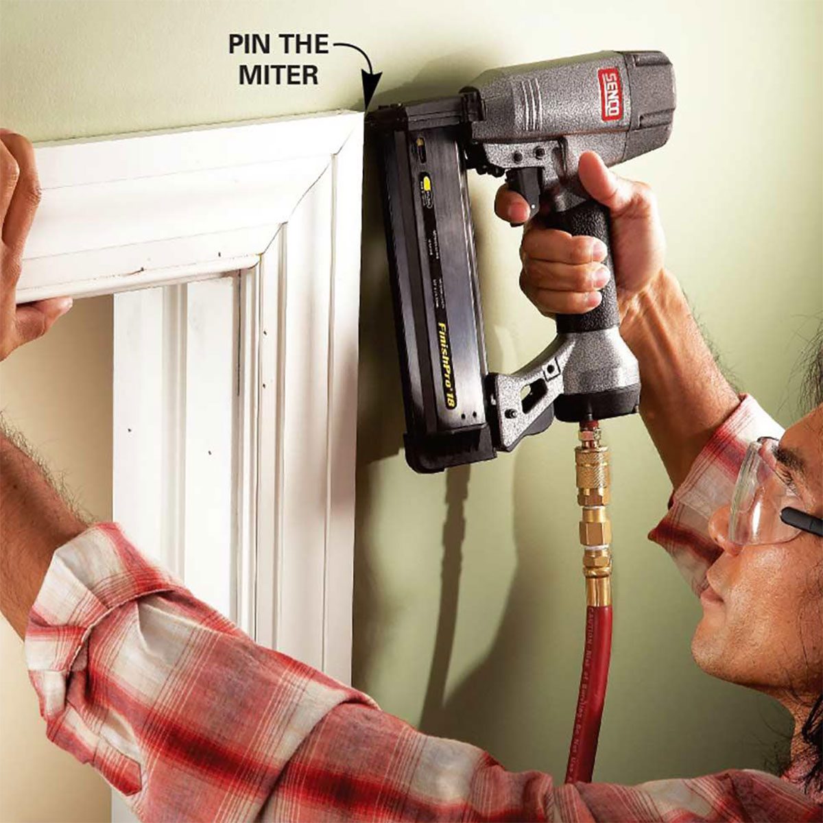 A person uses a nail gun to secure a miter joint in molding against a green wall, ensuring accuracy in the installation process.