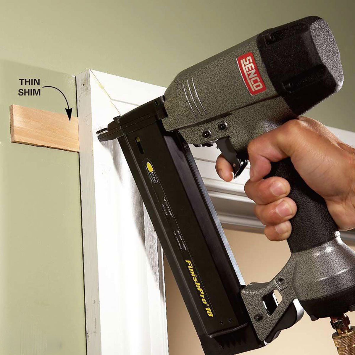 A person is using a nail gun to fasten a thin shim against a wall, surrounded by painted surfaces and a door frame.