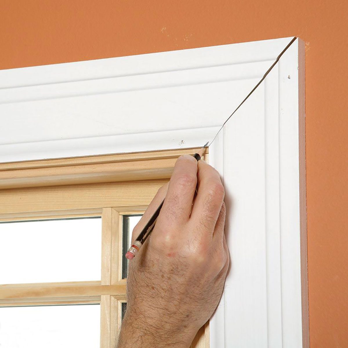 A hand holds a pencil, marking a white window trim near an orange wall, indicating preparation for adjustments or measurements.