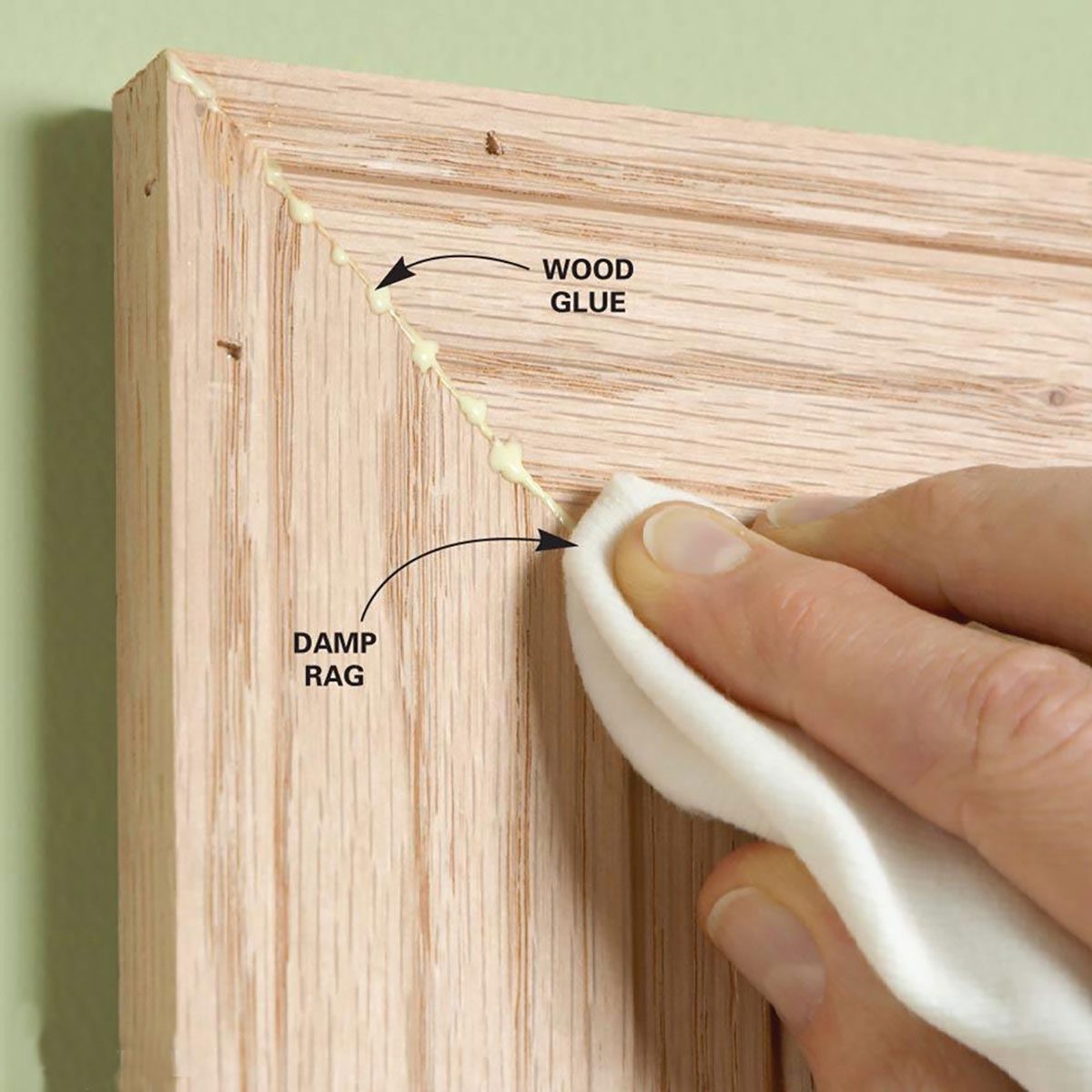 A hand wipes excess wood glue from a wooden surface using a damp rag, with a light green wall in the background.
