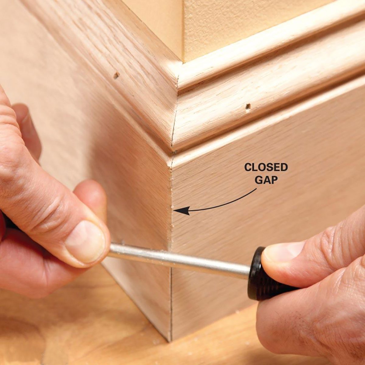 A hand uses a screwdriver to tighten a joint, closing a gap between two wooden pieces at the corner of a room.