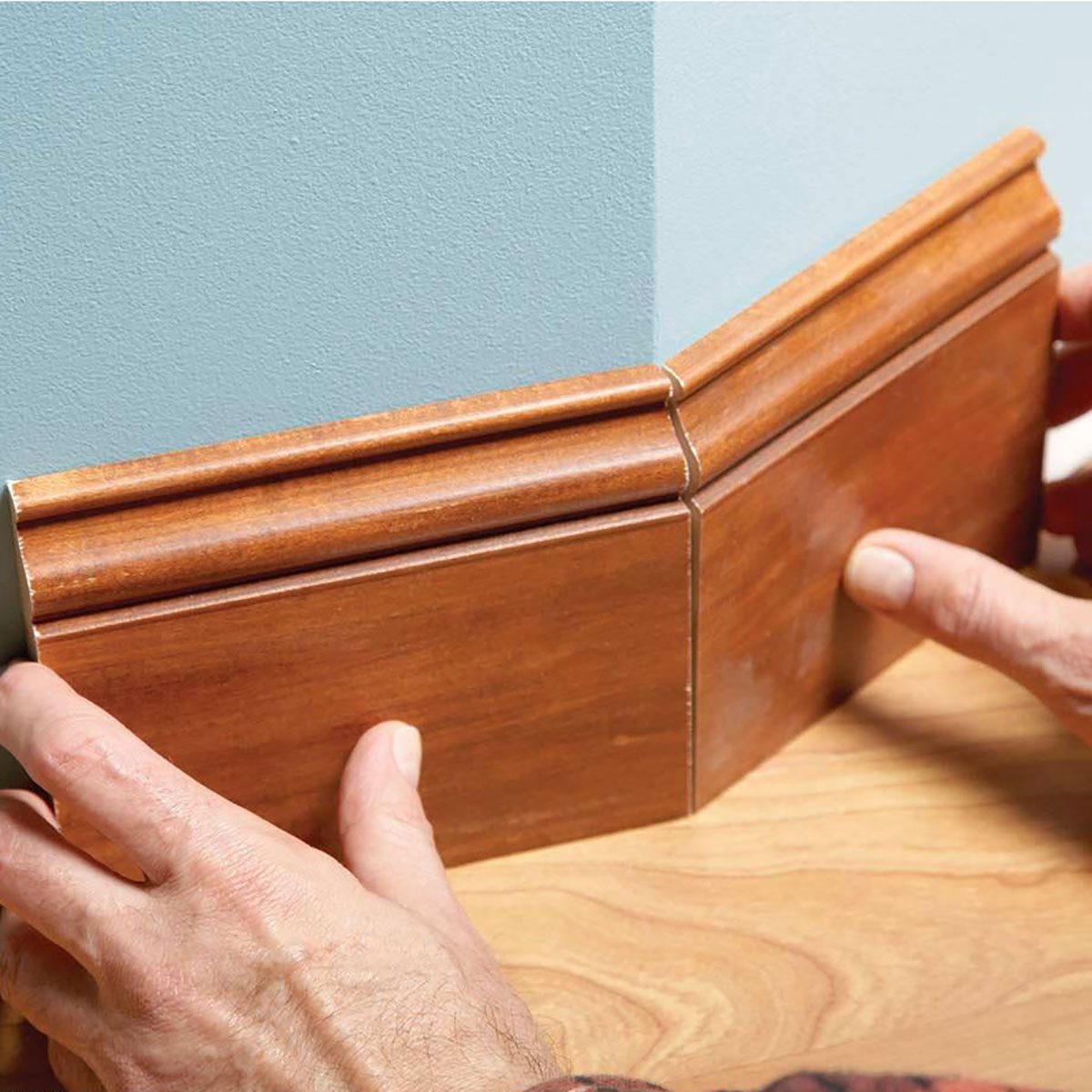 A person's hands are positioning a wooden corner molding piece against a wall, aligning it with a light blue surface in a wooden floor setting.