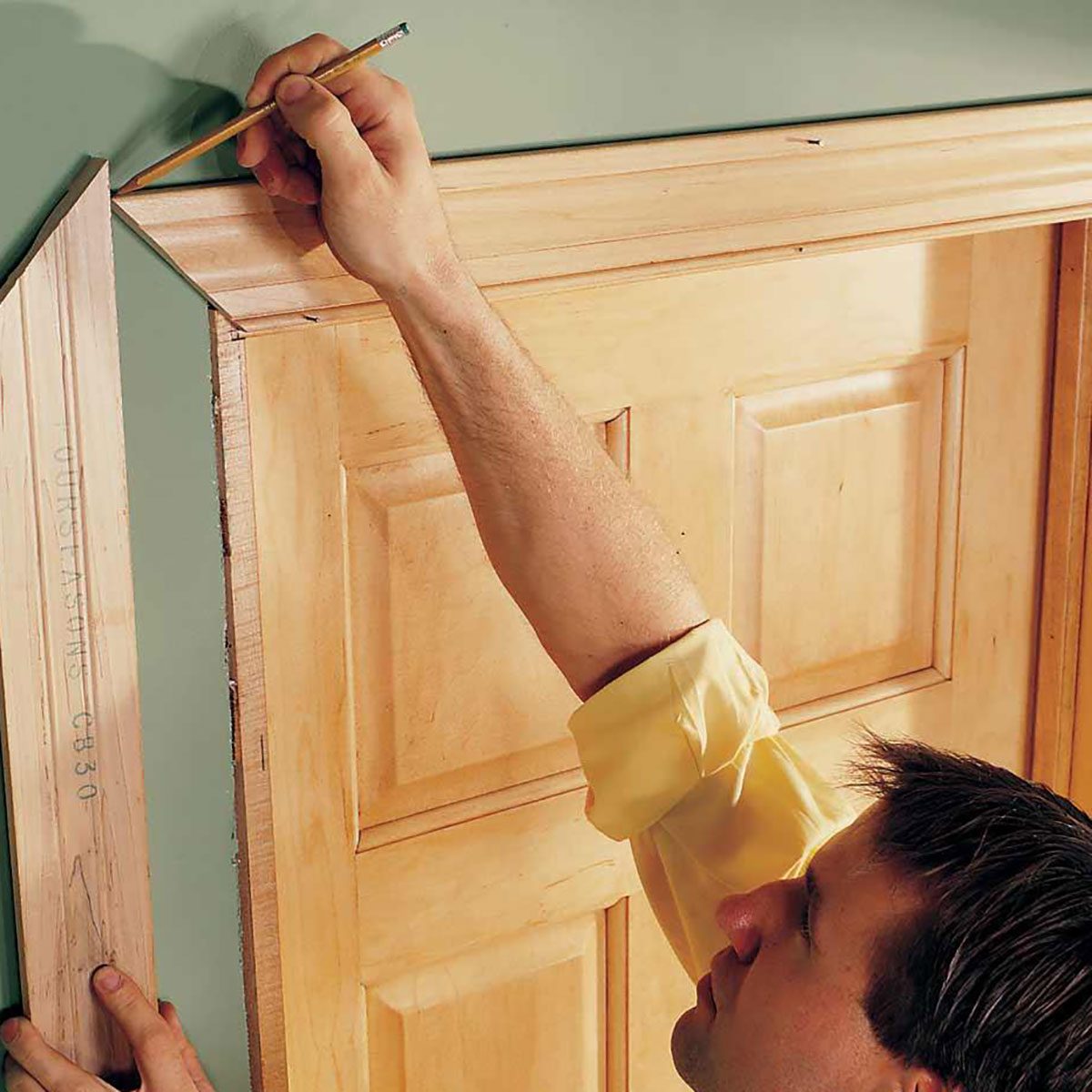A person holds a pencil, marking the corner of a wooden door frame against a green wall, preparing for an installation task.