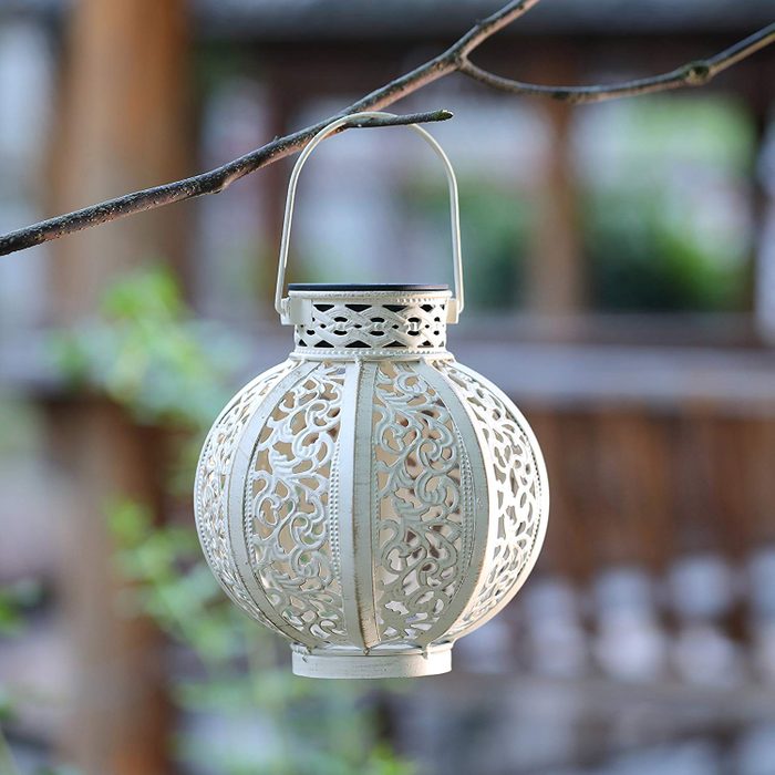 A decorative lantern hangs from a branch, showcasing intricate cut-out designs, suspended in a tranquil setting with blurred natural elements in the background.