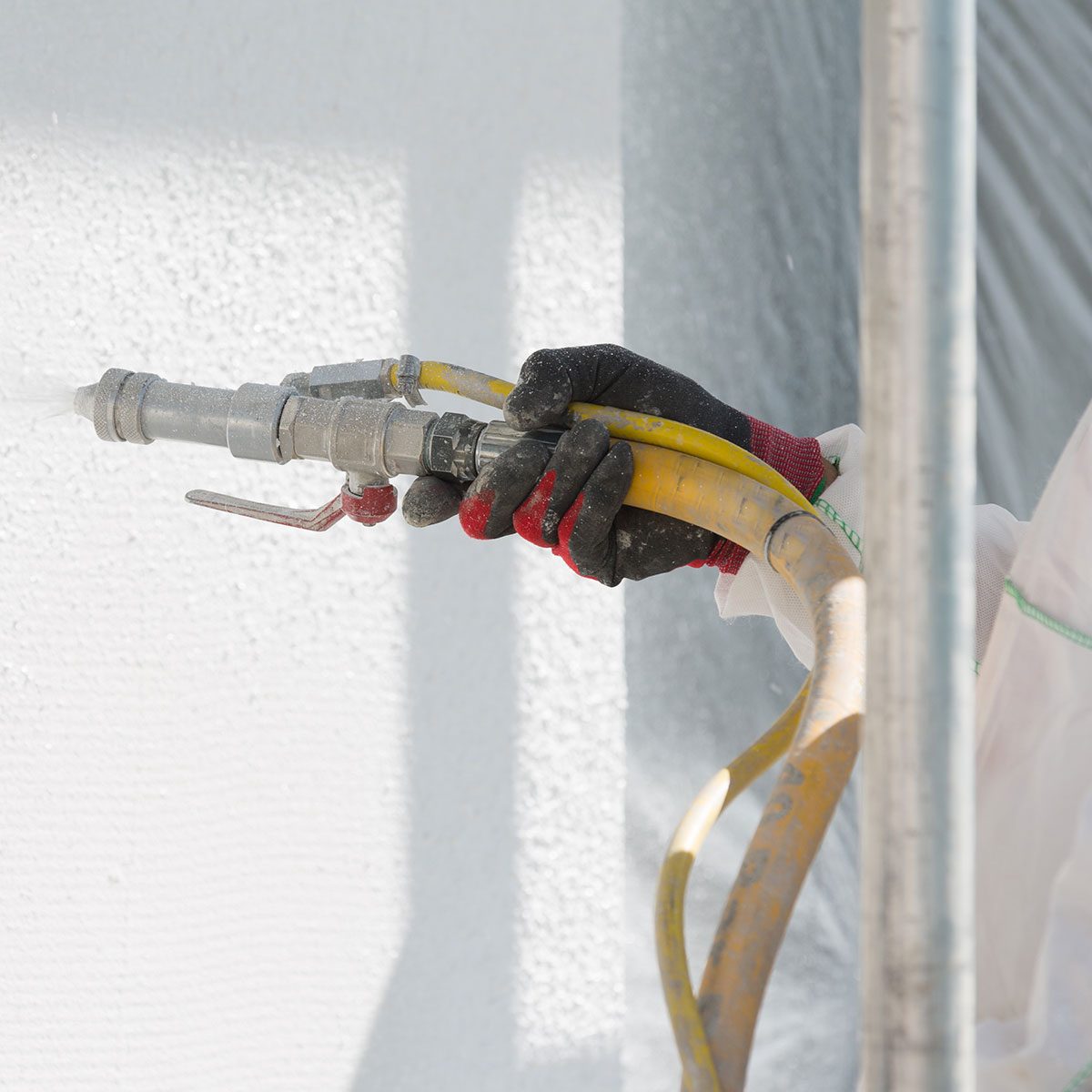 A gloved hand holds a spray nozzle connected to a yellow hose, while spraying a white textured wall in a brightly lit, construction environment.