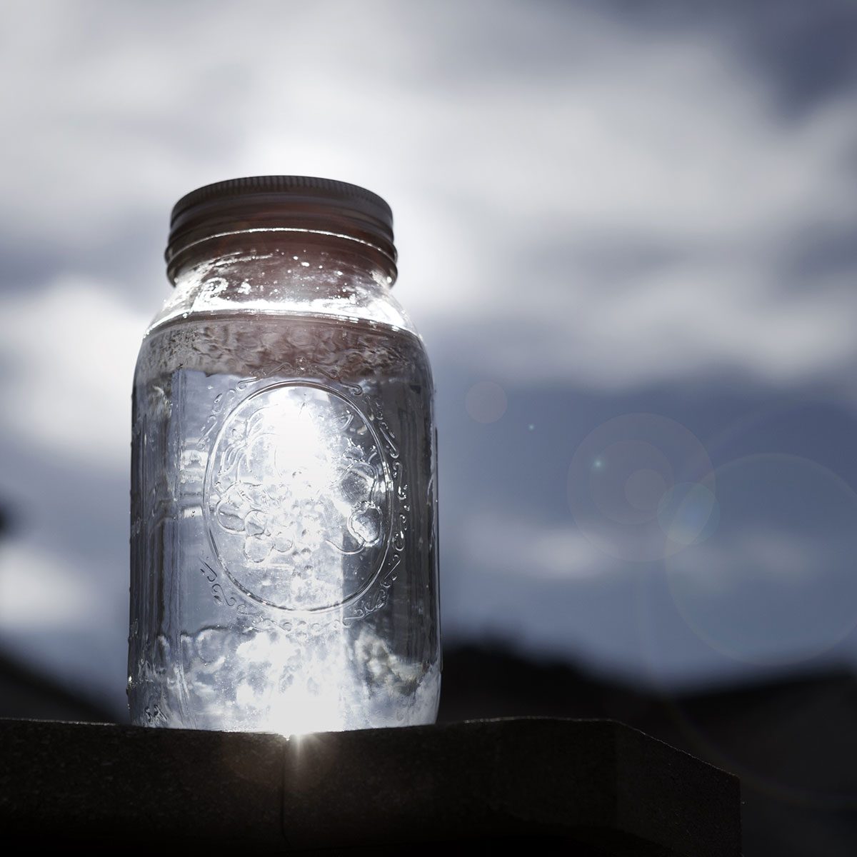 A glass jar filled with water reflects sunlight, sitting on a surface against a backdrop of cloudy skies.