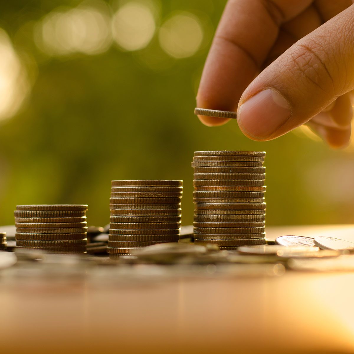 A hand hovers above three stacks of coins, adding a coin to the tallest stack while other coins scatter on a surface, with a blurred green background.