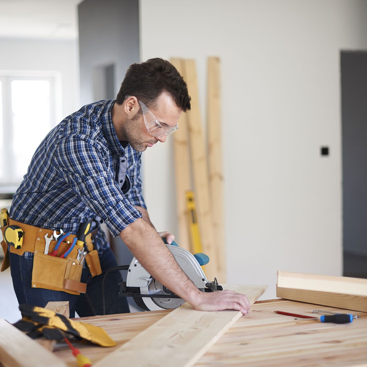 A man uses a circular saw to cut a wooden plank on a table, surrounded by tools, with wooden beams in the background of a well-lit room.