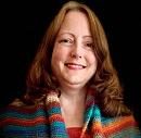 A woman smiles while wearing a colorful, patterned shawl. She is set against a dark background that accentuates her features.