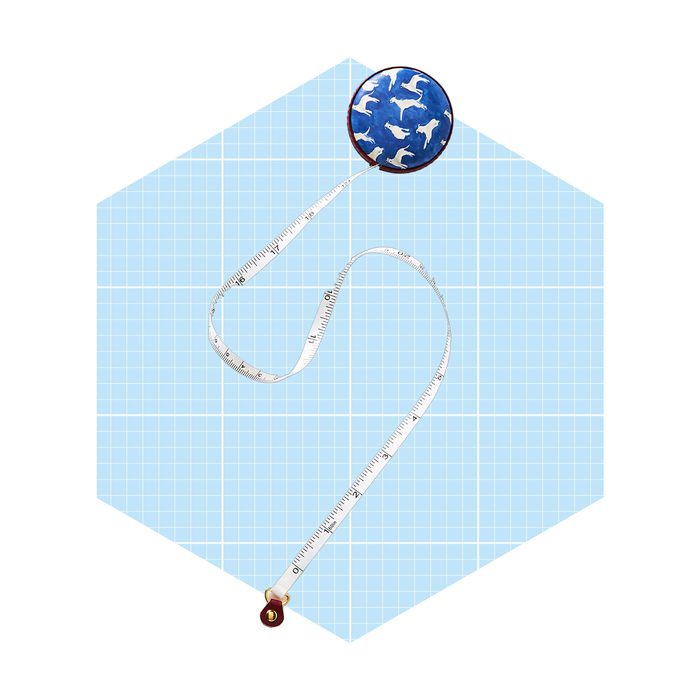 A measuring tape is extended with a patterned round case, lying on a light blue grid background, suggesting a crafting or design context.